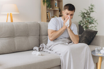 Sick man suffering from fever and headache blowing his nose into a napkin while sitting on the...