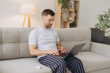 One man caucasian male freelancer work from home with headphones on his head on laptop computer customer support dispatcher while sit on sofa bed at home in shirt and pajamas happy smile