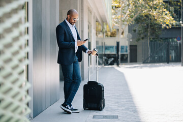 Businessman standing with suitcase and using mobile phone on sunny day