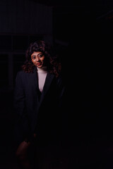 A woman in a black jacket and white sweater stands in a dark room. She is smiling and looking at the camera