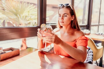 woman in a cafe seated, holding beverage. Dressed in an orange dress with her hair down. Bright interior, large windows allow natural light. Beverage consumption for refreshment.