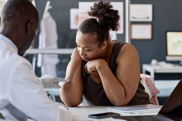 Dermatology clinic scene with doctor pointing at skin rash on arm of female patient listening to...