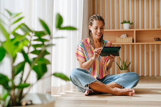 Happy young woman using tablet PC and sitting near window on floor