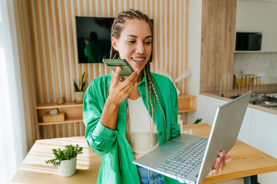 Happy young freelancer sending voicemail through smart phone and using laptop at home