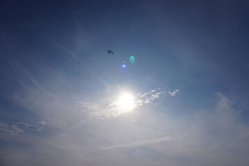The airplane is flying in the sunny blue sky and the sun is shining behind the white clouds 