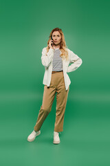 A confident woman with curly hair chats on her phone, dressed casually in a stylish outfit against a green backdrop.