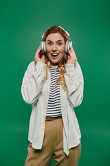 A lively woman with curly hair expresses joy while wearing headphones on a vivid green background.