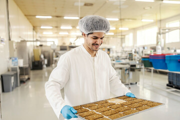 Smiling interracial food factory worker carrying tray with baked biscuits at food processing plant.