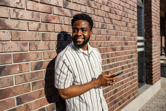 Happy black man standing against the wall at train station and typing messages while looking away.