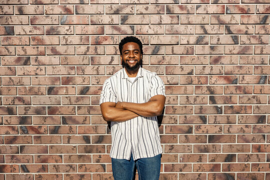 Portrait of a young smiling black guy standing near brick wall with arms crossed.