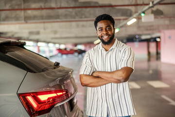 Portrait of a smiling black man standing next to his car in public garage with arms crossed.