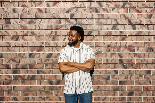 Portrait of an african american casual man standing with arms crossed near brick wall and looking away