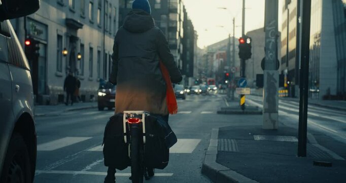 Woman in winter clothing on cargo electric bicycle wait on traffic light. Cyclist riding with car traffic, crossing tram lines on bike. Cyclist city commuting in early morning on the way to office