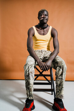 man with vitiligo wears a yellow tank top and camouflage pants, sitting on a stool facing the camera with a composed expression, against a brown background, reflecting strength and individuality