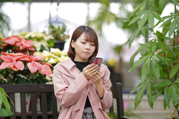A young woman in her 20s wearing a pink coat is sitting on a bench in front of white and pink poinsettias and operating a smartphone.