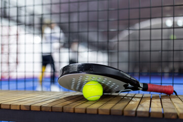 A close-up of a paddle and ball resting on a wooden bench during a padel match in an indoor court...