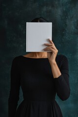 Naklejka premium A woman in a black dress holds a blank book in front of her face against a dark green background. She is shrouded in mystery and intrigue.