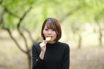 A young woman in her 20s with short hair wearing a checked skirt and black knit long sleeves is sitting on a bench in the forest and eating rusk with a delicious smile.