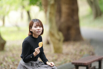 A young woman in her 20s with short hair wearing a checked skirt and black knit long sleeves is sitting on a bench in the forest and eating rusk with a delicious smile.