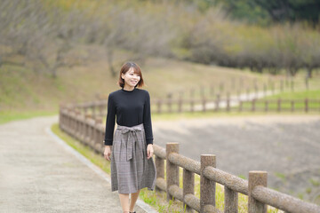 A young woman in her 20s with short hair wearing a black knitted long-sleeved shirt is walking alone on a path along a lake in the forest.