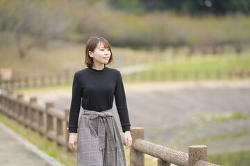 A young woman in her 20s with short hair wearing a black knitted long-sleeved shirt is walking alone on a path along a lake in the forest.