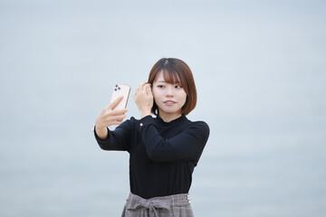 A young woman in her 20s with short hair wearing a black knitted long-sleeved shirt is taking a selfie with a smartphone on the sandy beach by the sea.