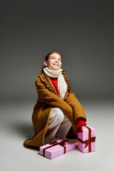 A young girl in warm attire smiles happily while hugging a blanket and surrounded by gifts