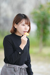 Fototapeta premium A young woman in her 20s with short hair wearing a black knitted long-sleeved shirt is sitting in a pavilion in an empty park and eating a cookie with a delicious smile.