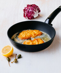 Two breaded Schnitzel sizzling in a black pan on a white surface. There is a lemon, capers, and a...