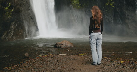 Tourist woman looking powerful Bali waterfall, water cascading down rocks. Long hair blows in wind....