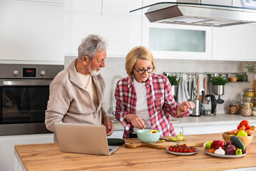 Senior couple man and woman cooking meal together in domestic kitchen at home using laptop computer for new recipe they find online on internet. Fun and respect, togetherness of retired couple.