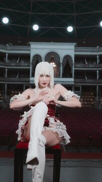 A stylish young woman in a red dress, white thigh highs, and an elaborate pearl headdress, sits on a chair on stage and dances alluringly in an empty theater.