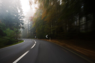 Fototapeta premium Long exposure, blurry autumn morning covered empty rural road through a forest in Europe. Wide angle view, limited visibility, bad driving weather concept, no people