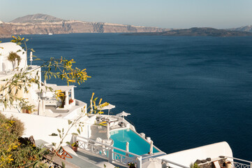 Panoramic view from the top of the Oia caldera on the island of Santorini in Greece. White buildings, terraces and pools overlook the blue waters of the Aegean Sea, surrounded by jagged cliffs.
