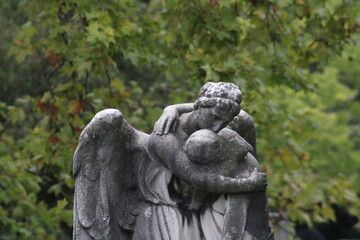 A solemn stone angel embraces a figure in a tranquil cemetery surrounded by lush greenery during an...