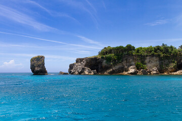 Gentle waves lap against rugged cliffs and a towering rock formation under a vibrant blue sky. Lush...