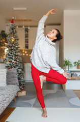 Woman stands in living room and doing yoga against background of Christmas tree.