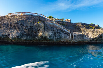 Stone staircases wind along the edge of a rocky cliff, overlooking a body of clear blue water....