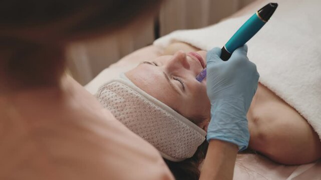 Expert cosmetologist uses a derma pen for facial microneedling therapy on a relaxed client lying down in a wellness spa.