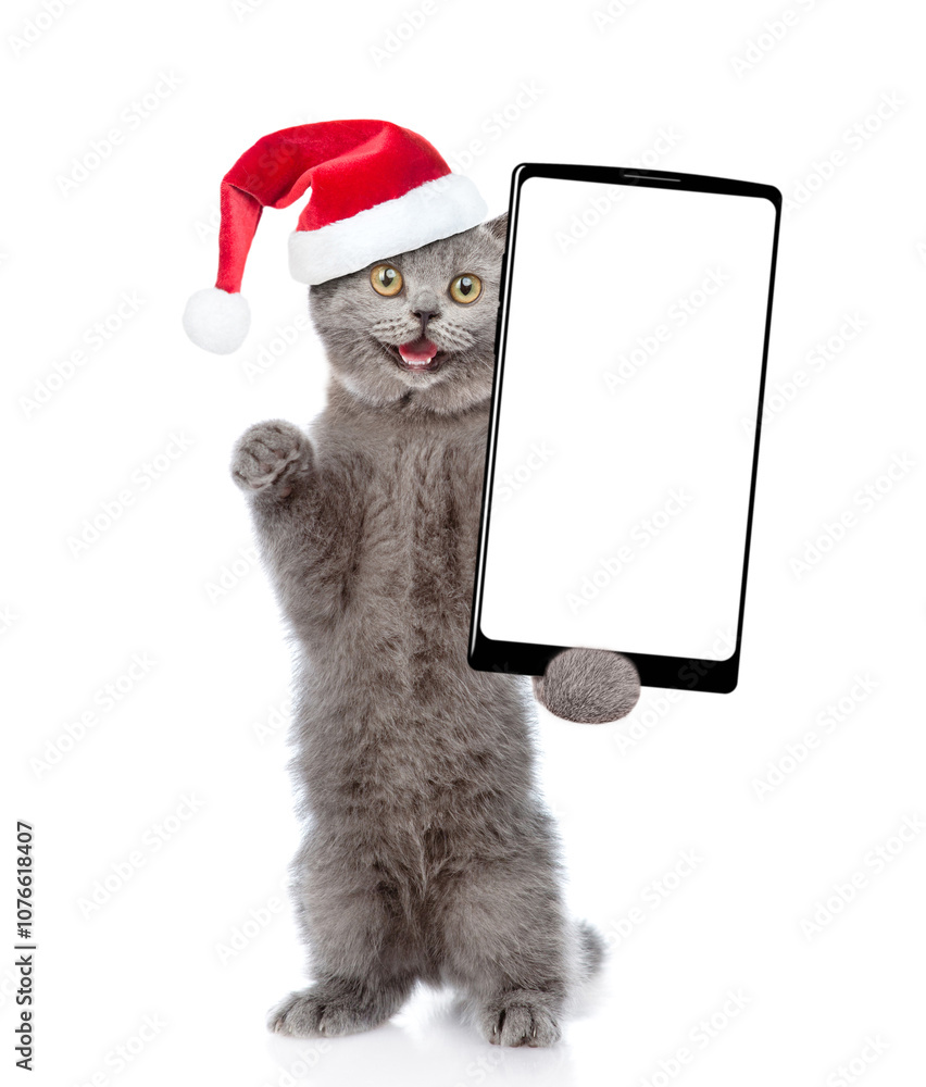 Poster Surprised kitten wearing santa hat holds smartphone with white blank screen in it paw. Empty free space for mock up