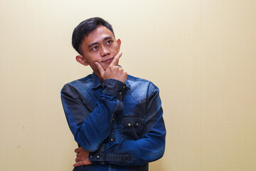 A young Asian man in a blue shirt stands thinking with his hand on his chin and arms crossed, on a...