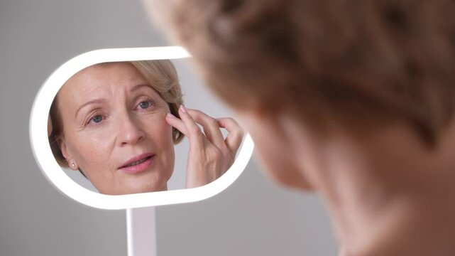 A mature blond woman examines her skin, sad about wrinkles and imperfections. Anti-aging skincare routine, graceful aging and selflove.