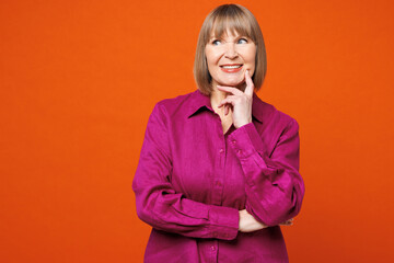 Elderly blonde woman 50s years old wear purple shirt casual clothes put hand prop up on chin, lost in thought and conjectures isolated on plain red orange background studio portrait Lifestyle concept