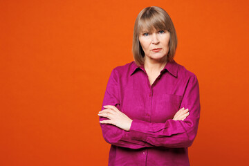 Elderly sad mad frowning blonde woman 50s years old wear purple shirt casual clothes hold hands crossed folded look camera isolated on plain red orange background studio portrait. Lifestyle concept.