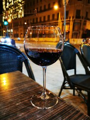 A glass of red wine on a table in a bustling outdoor café at night, with city lights creating a vibrant atmosphere