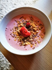 Bowls of vibrant pink smoothie topped with granola, flaxseeds, and raspberries placed on a wooden table at breakfast time