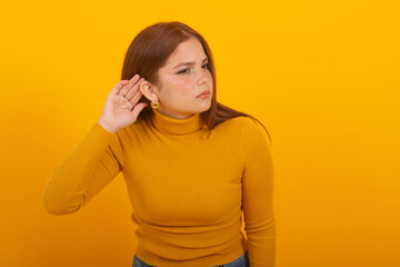 Frustrated Young Redhead woman wearing a yellow sweater Struggling to Listen hand behind ear on a...