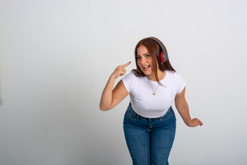 Horizontal full-body portrait of a young pretty redhead lady listening to music enjoying and pointing to her headphones isolated on a white background