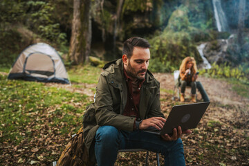 Fototapeta premium adult man hiker work remote on laptop in front waterfall and tent