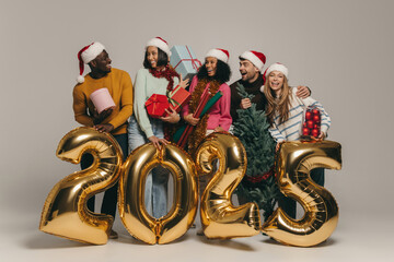 Joyful friends holding Christmas ornaments and gift boxes while celebrating 2025 New Year on studio background